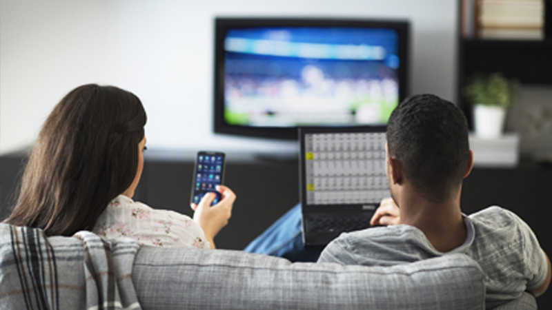 A woman using a smartphone and man using a laptop both sitting on a couch in front of a television.