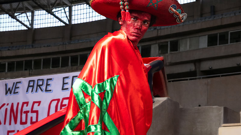 Person wearing a red hat and cape adorned with a green star, reminiscent of Morocco's flag. Their face is painted red. A stadium is in the background.