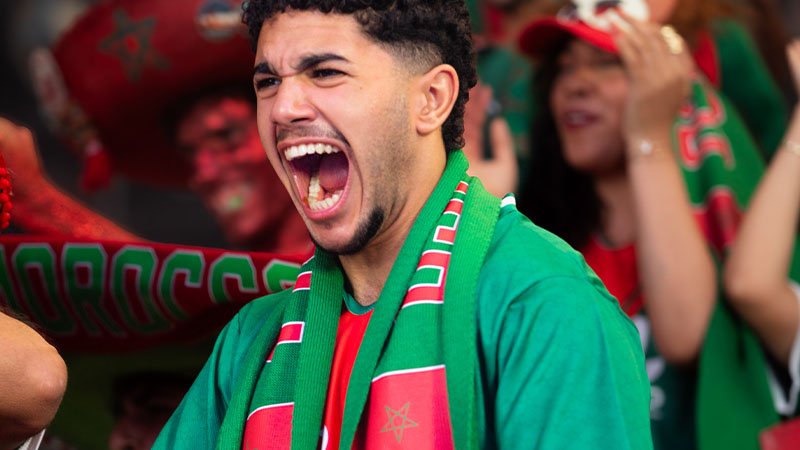 A passionate fan wearing a Moroccan flag-themed scarf and jersey cheers energetically, surrounded by fellow supporters in vibrant green and red attire.