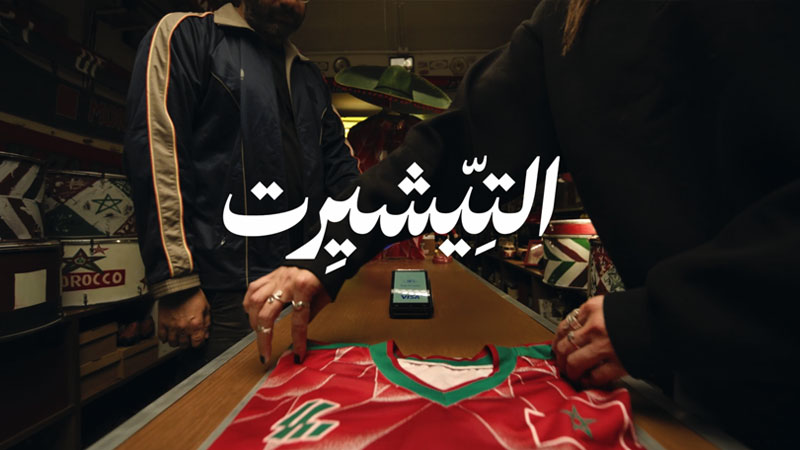 Two people standing over a counter in a dimly lit shop, exchanging a red sports jersey. A smartphone and a Visa card are placed on the counter.