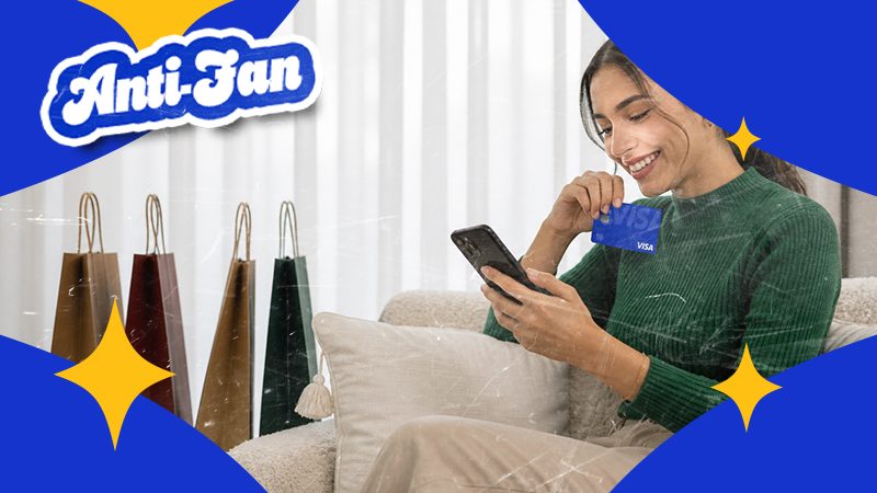 Smiling woman in a green sweater holds a Visa card and smartphone, sitting on a couch. Shopping bags beside her, with "Anti-Fan" text above.